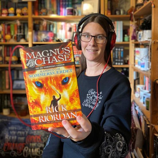 Line sidder foran en fyldt bogreol. I hånden har hun en bog, med en høretelefonledning i, der fører op til det headset hun har på.