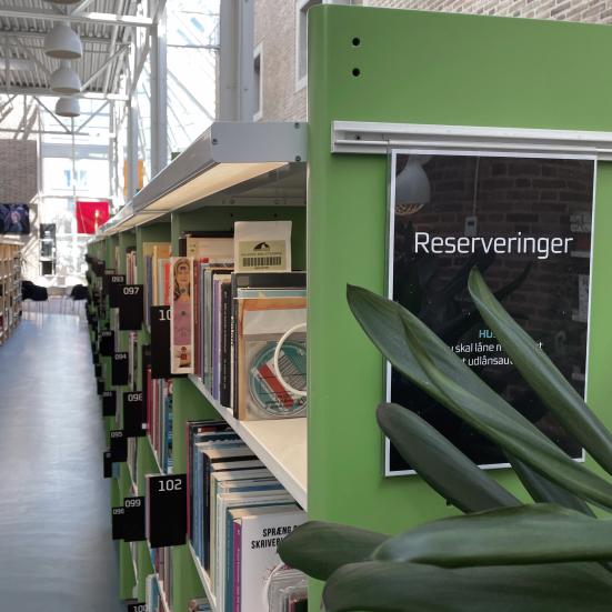 De grønne reserveringshylder på Hovedbiblioteket. I forgrunden bag en plante står et skilt med "Rerserveringer" skrevet på.