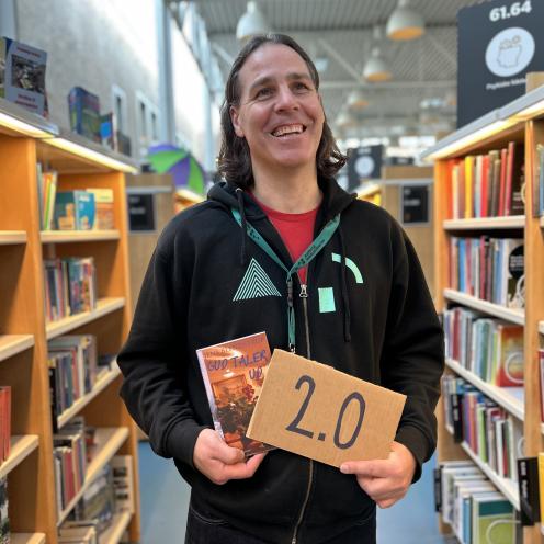 Kenneth står blandt fyldte bogreoler på Hovedbiblioteket. Han holder romanen "Gud taler ud" af Jens Blendstrups i den ene hånd og et brunt papskilt hvorpå der står "2.0". Kenneth kigger smilende ud i biblioteksrummet.