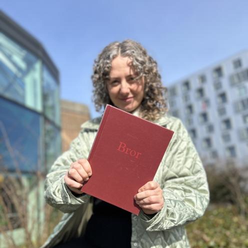 Majken står i bibliotekshaven ved Hovebiblioteket. Hun holder bogen "Bror" frem for sig og smiler. Både Majken og baggrunden er let sløret, så der er fokus på bogforsiden. Himlen er blå.