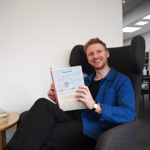 Patrick sidder i en lænestol og læser i "En linje i verden". Bag ham ses biblioteksrummet. Han kigger ind i kameraet og smiler. 