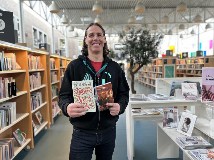 Kenneth står på Hovedbiblioteket. Han holder to af Jens Blendstrups romaner foran sig og smiler til kameraet. Bag Kenneth ses biblioteksrummet med fyldte bogreoler og bogudstillinger.
