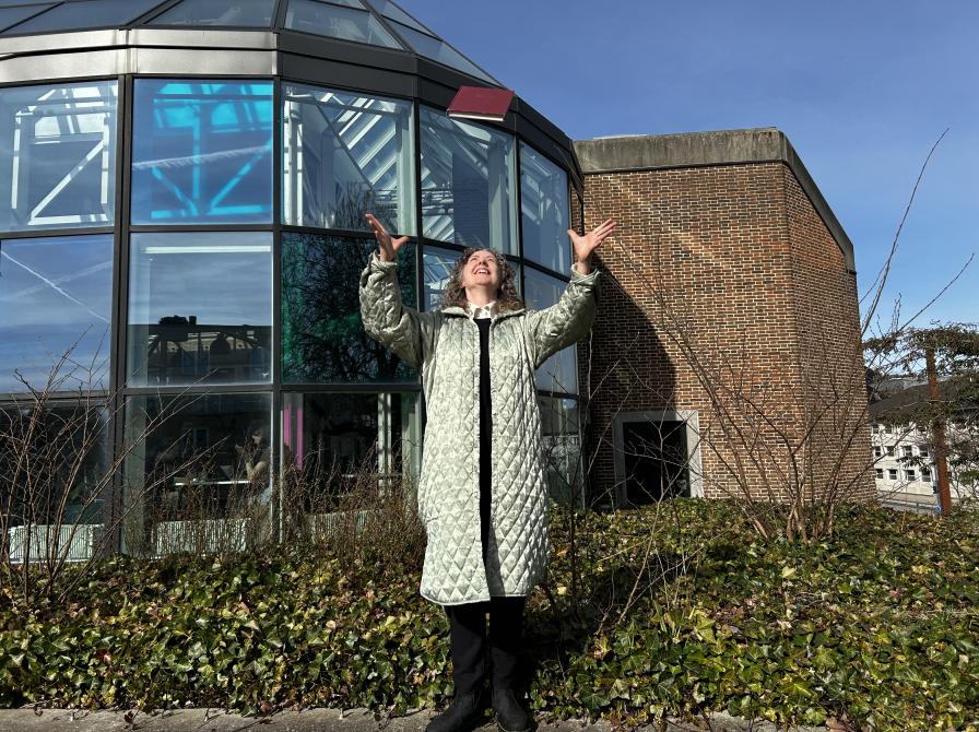 Majken står i bibliotekshaven ved Hovebiblioteket. Hun kaster bogen "Bror" op i luften og rækker hænderne op for at gribe den igen. Majken smiler og bag hendes ses glashjørnets farverige vinduer. Himlen er blå.