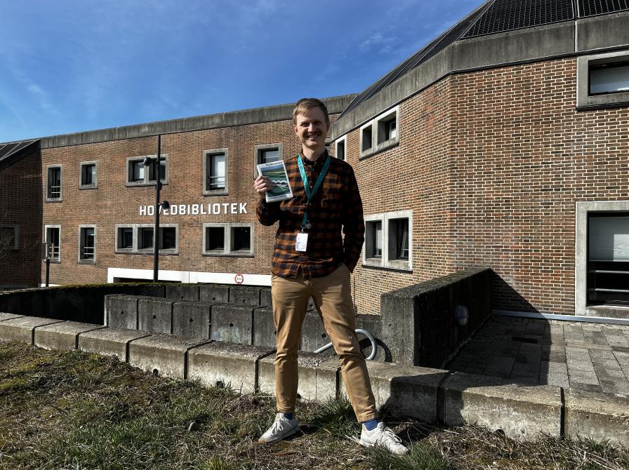 Mathias står i bibliotekshaven ved Hovedbiblioteket. Han holder bogen "Hyldemeter" i den ene hånd og har den anden hånd i lommen. Mathias smiler til kameraet. Himlen er blå og solen skinner.